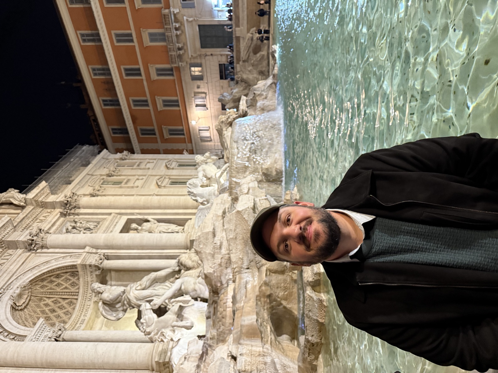 Gabis de boné sorrindo em frente à Fontana di Trevi, em Roma, à noite