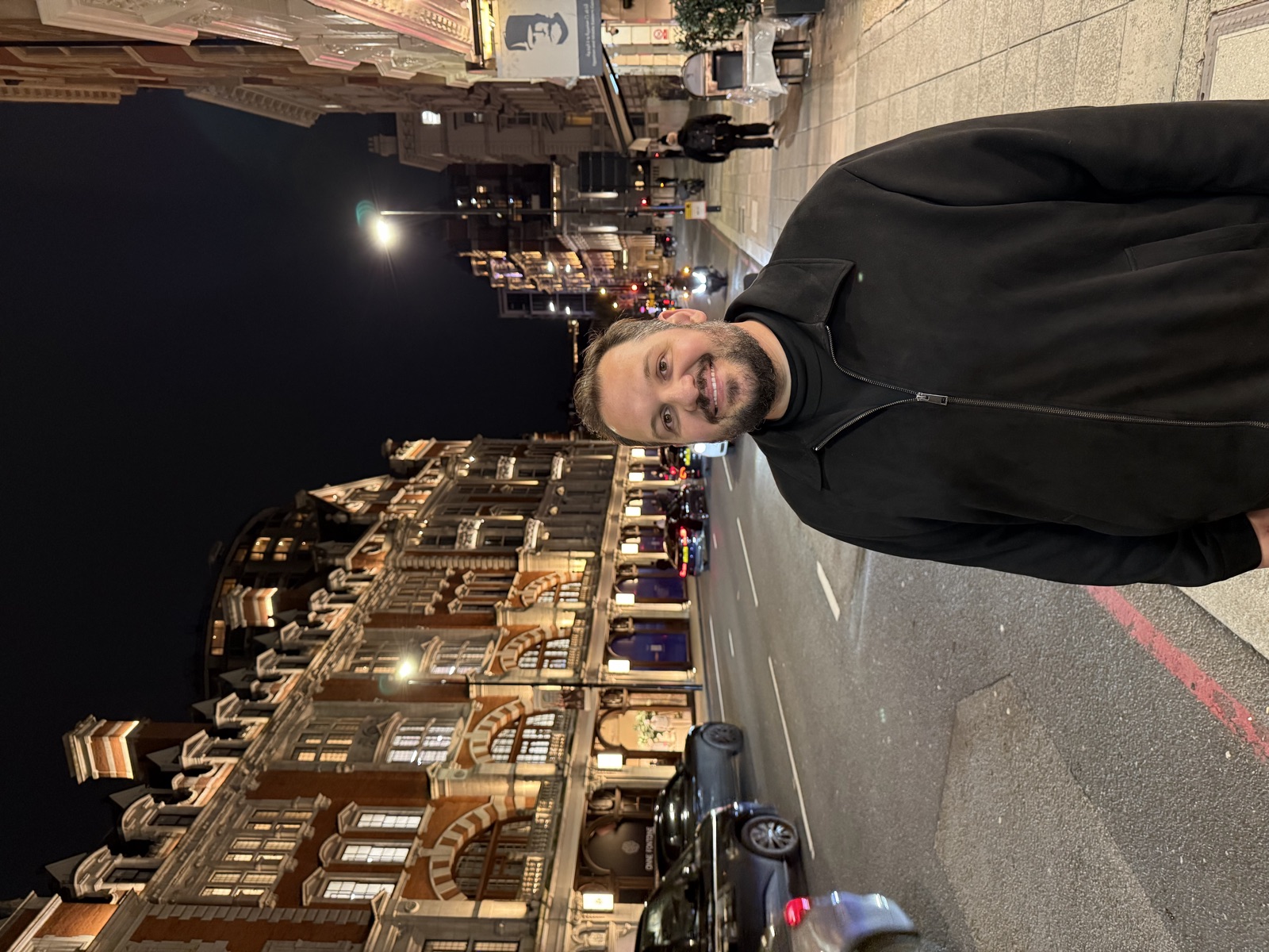 Gabis sorrindo em uma avenida de Londres à noite, com prédios históricos iluminados ao fundo